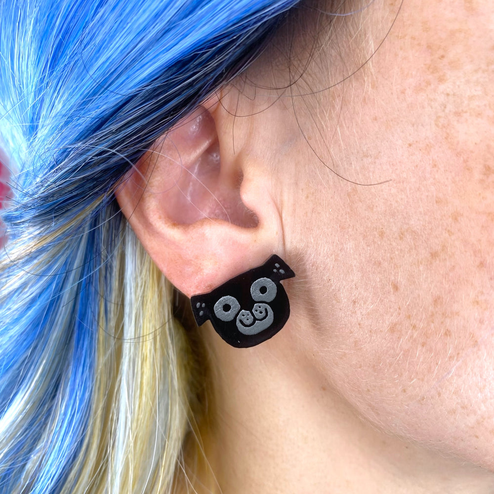 A black pug stud earring illustrated by Gemma Correll and worn by a person with bright blue hair.