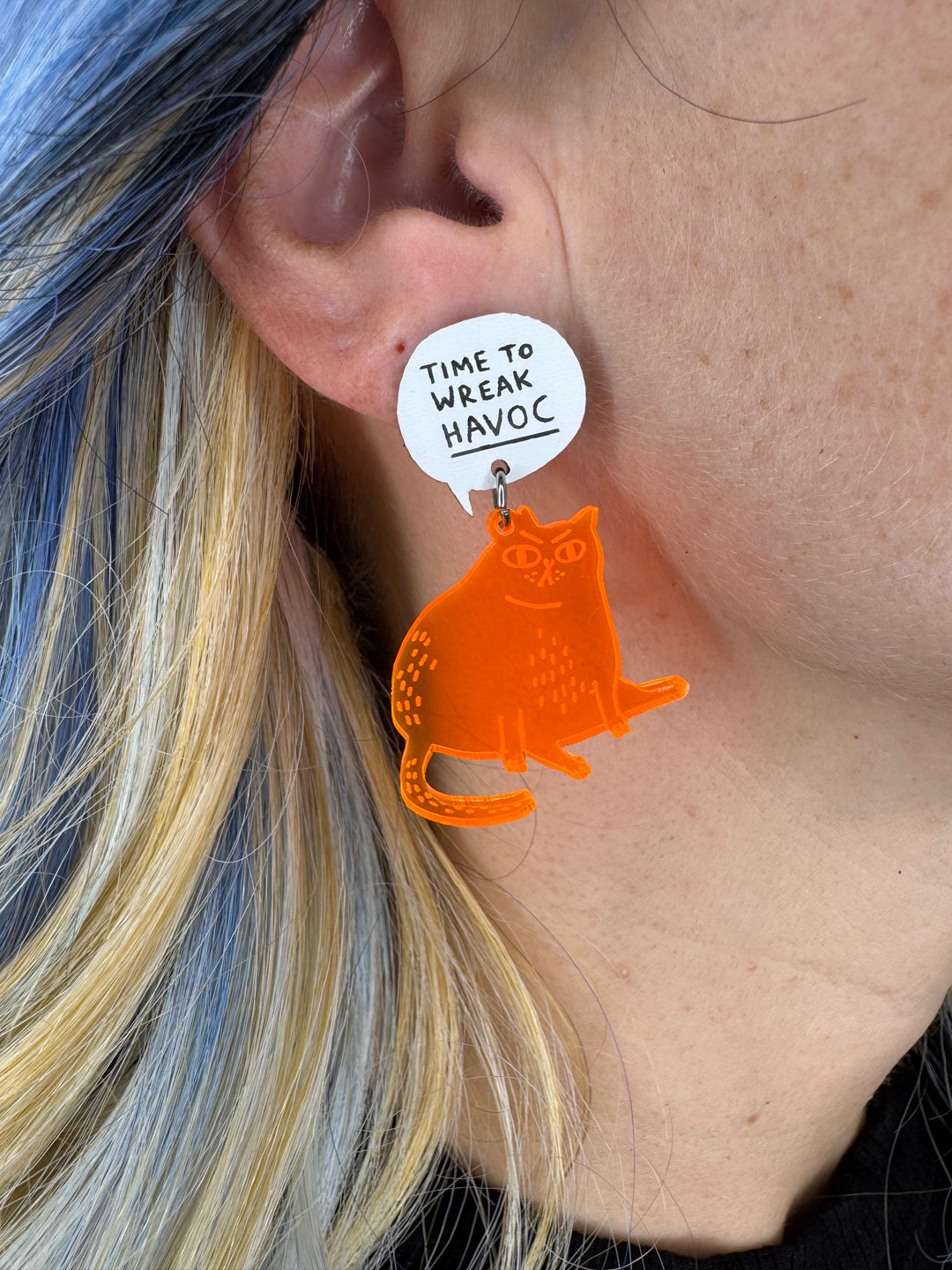 Transparent orange cat earring with a white tag worn by a person with blue and blonde hair.