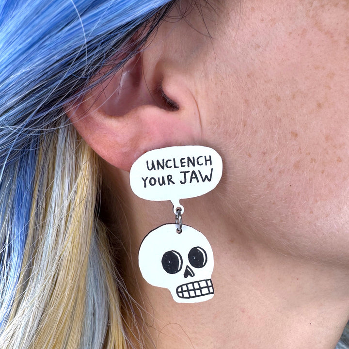 Gemma Correll skull-shaped earring with 'Unclench Your Jaw' speech bubble worn by a person with blue hair.