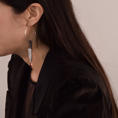 A woman with long brown hair wears a stainless steel hoop earring with plastic chef's knife charm and Austrian crystal blood drop.