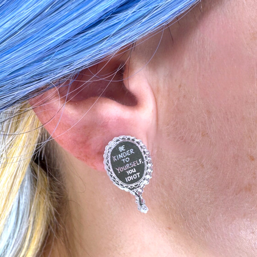 Earring with 'Be kinder to yourself, you idiot' text worn by a person with blue hair.
