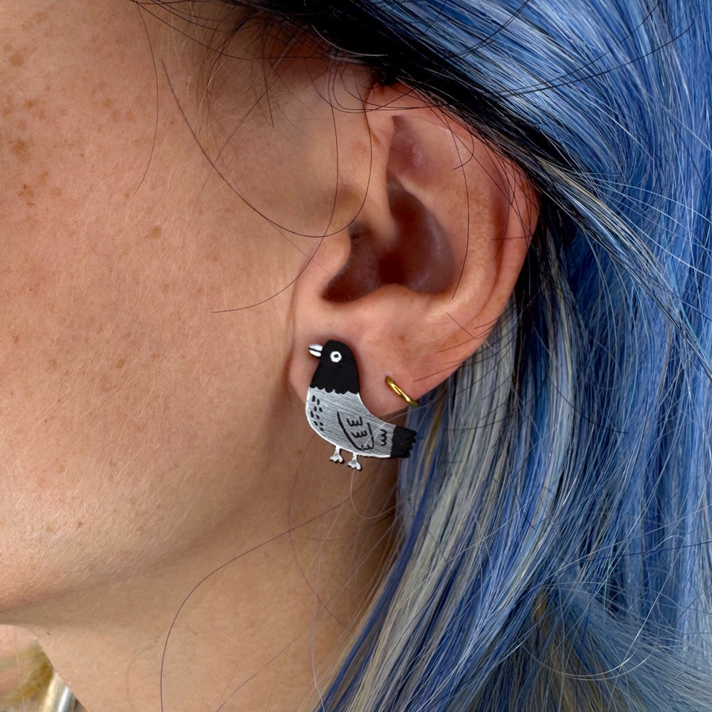 Close-up of a person with blue hair wearing Gemma Correll's Pigeon stud earring in brushed silver tone and matte black.