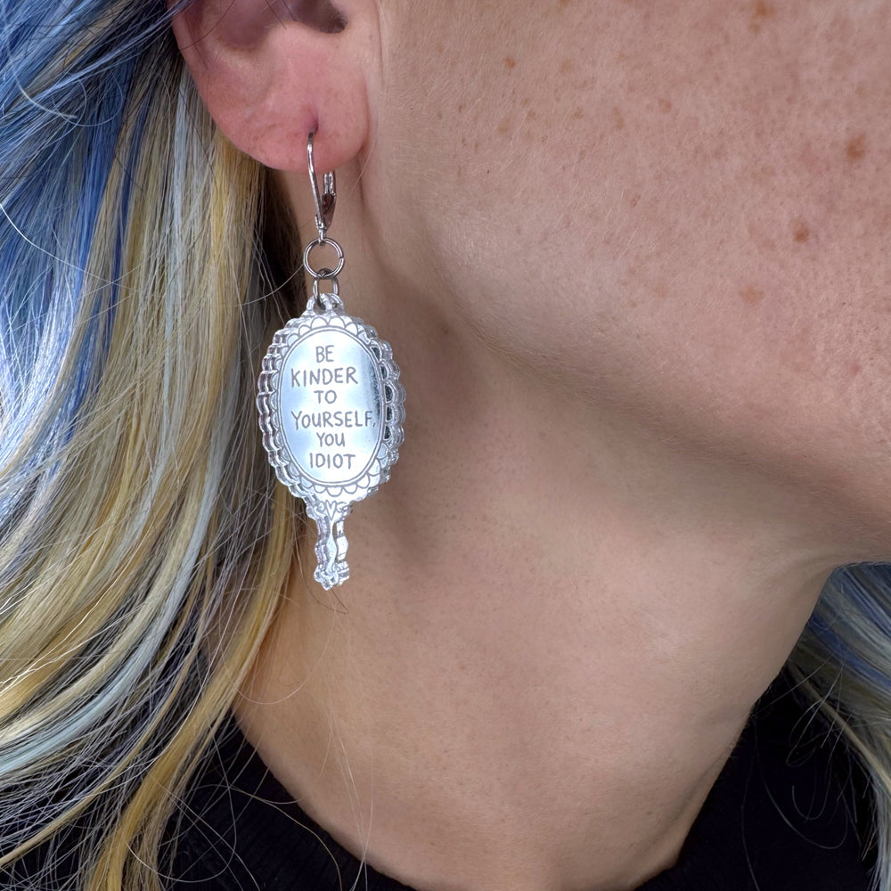 A person with blue/blonde hair is wearing a mirror dangle earring with the snarky phrase, 'Be Kinder to Yourself, You Idiot' by Gemma Correll.