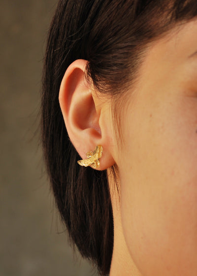 Mirror gold bee stud earring sits on a models earlobe with a taupe background.