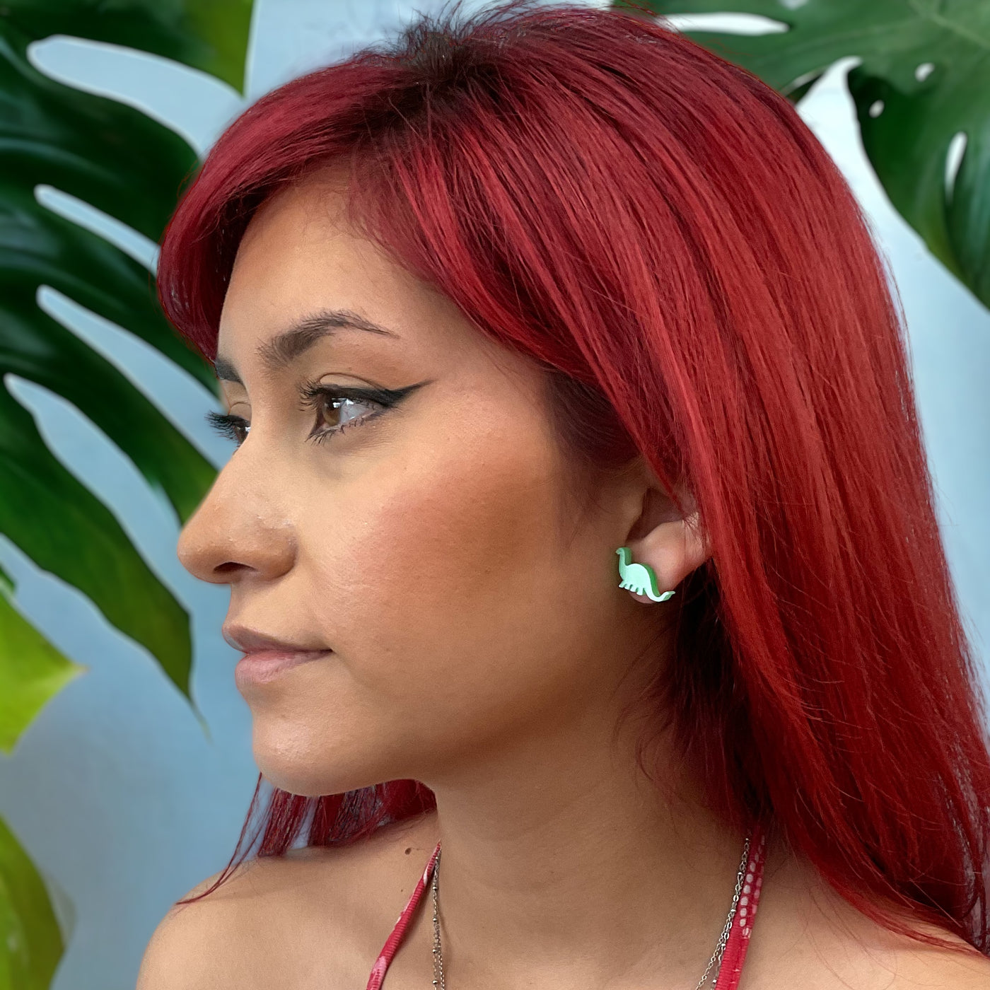 An alternatively styled model with bright red hair wears a plastic pearl green Brontosaurus dinosaur earring on her ear.