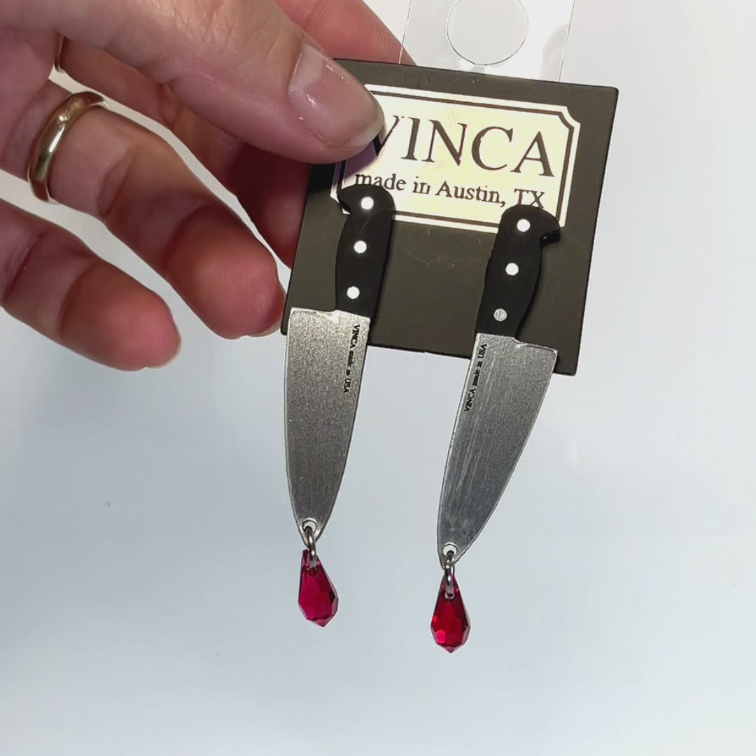 A hand showcases Vinca's Chef's Knife Earrings with Austrian crystal blood drop on a clack branded hang tag.