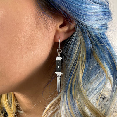 Blue and blonde haired woman wears silver tone switchblade dangle earring.