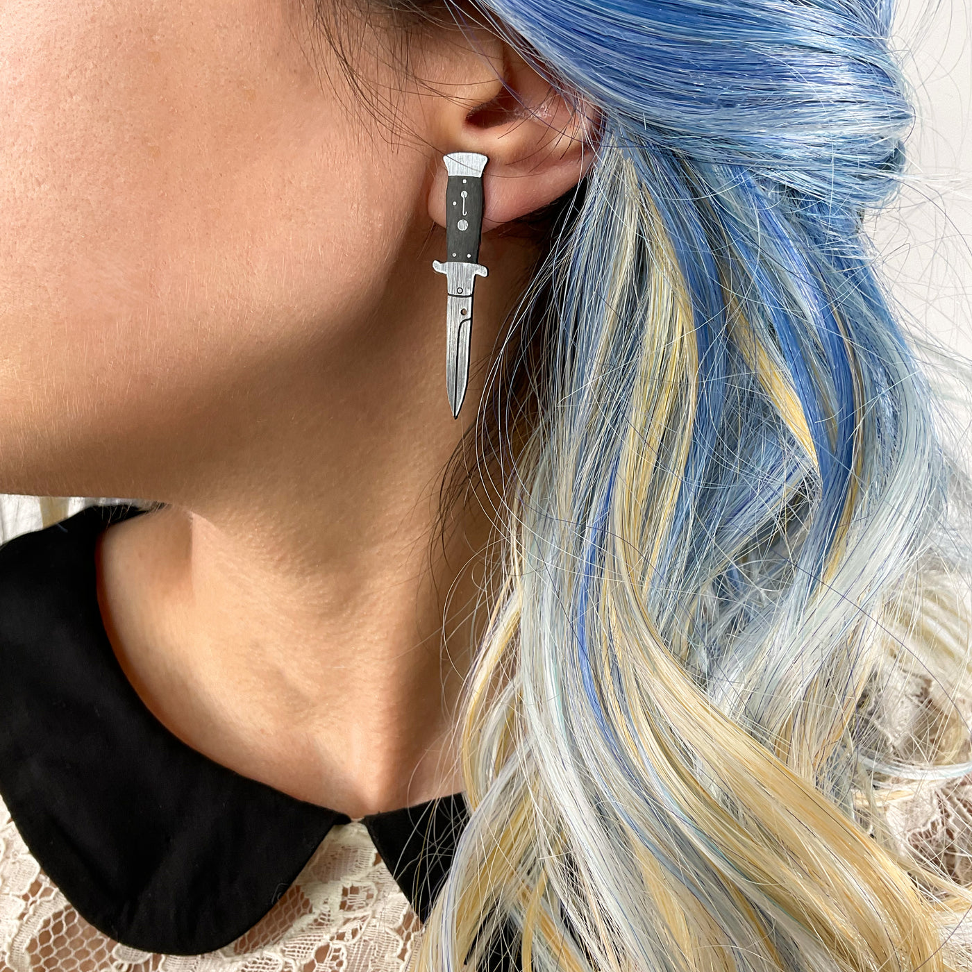 Silver switchblade stud earring on the earlobe of an alternative woman with blue hair with blonde streaks.