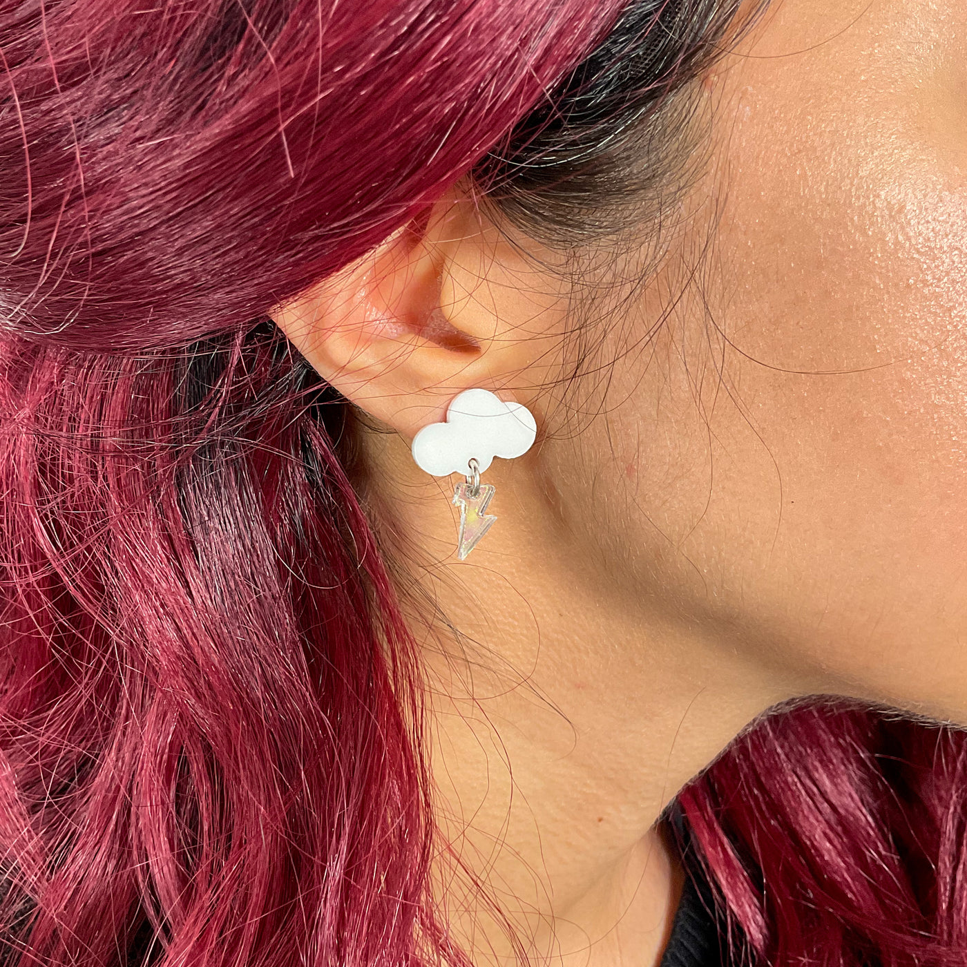 Solid white small cloud earrings with a transparent bolt on a woman's ear surrounded by burgundy hair. 