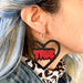 Person with colorful hair and a cheetah print top wears a black heart earring with red text that reads 