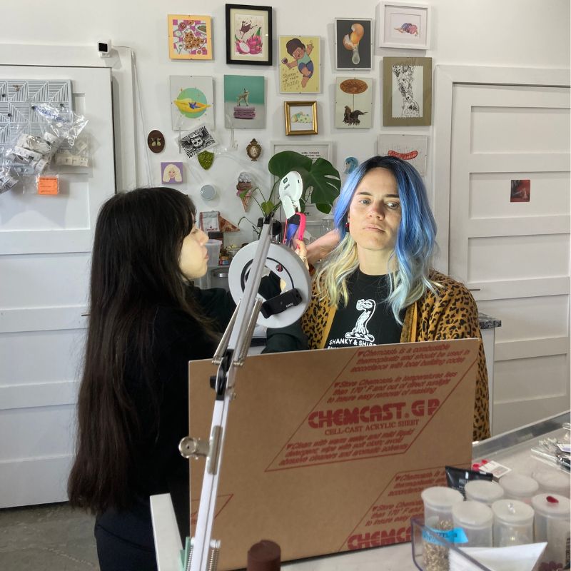 Two women that work at a Vinca, a small jewelry business, take product photos in house.