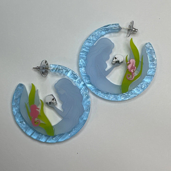 A pair of handmade hoop earrings made from light blue marbled acrylic, decorated with silver skull studs and green and pink floral accents, displayed on a neutral light gray background.