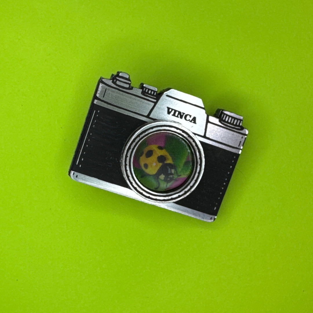 A vintage-style camera-shaped hairclip with a black textured body, silver metal edges, and a circular green-and-yellow panda lens detail, shown on a solid bright green background.