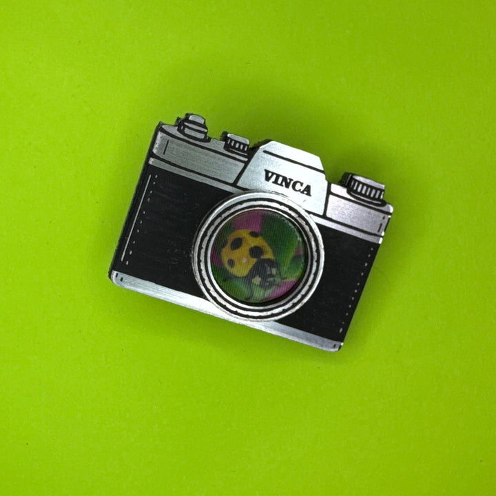 A vintage-style camera-shaped hairclip with a black textured body, silver metal edges, and a circular green-and-yellow panda lens detail, shown on a solid bright green background.