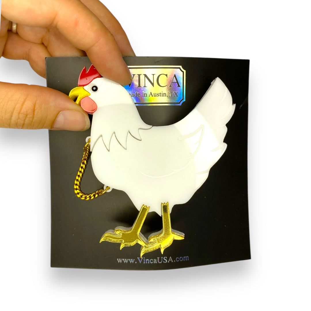 Our Chicken with its head cutoff brooch is shown on a black branded Vinca card with a hand pinched around the chicken's head.