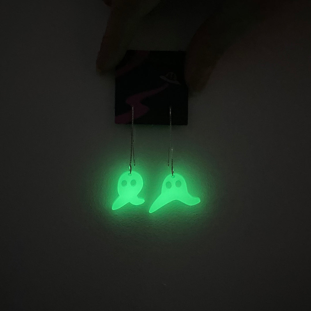 Sarah's Scribbles Cryptid Club Frenso Nightcrawler ear threader earrings glow in the dark against a white background in the dark.