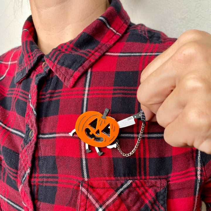 Person wearing a red and black plaid shirt with our interactive Halloween-themed Gemma Correll Jack-o' lantern brooch. 