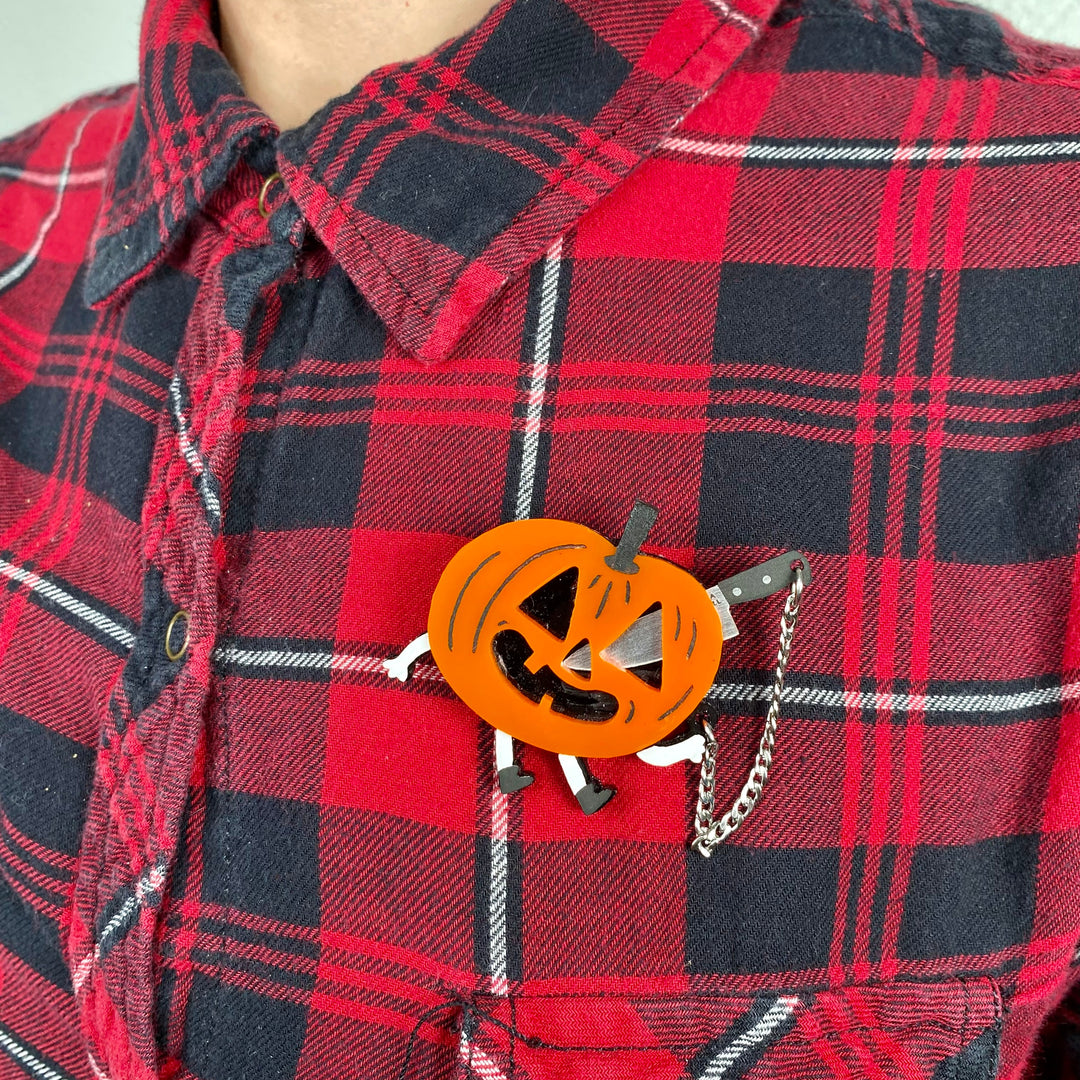 Our officially licensed Gemma Correll interactive Jack-o-lantern pumpkin brooch on a person wearing a red and black plaid shirt.