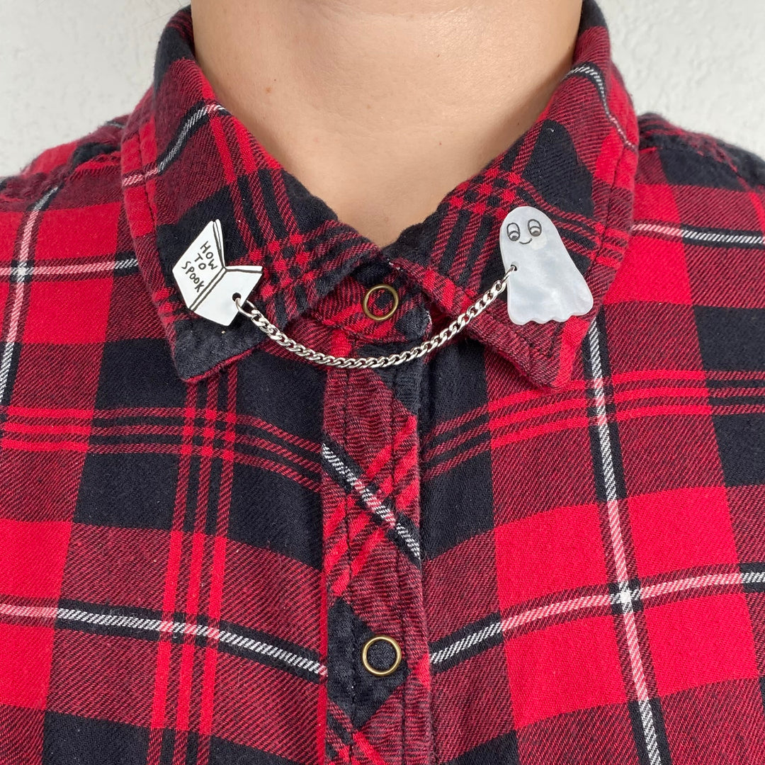 A person wears a red and black plaid shirt with our official licensed Gemma Correll Halloween-themed spooky ghost with 'How to' book collar clips.