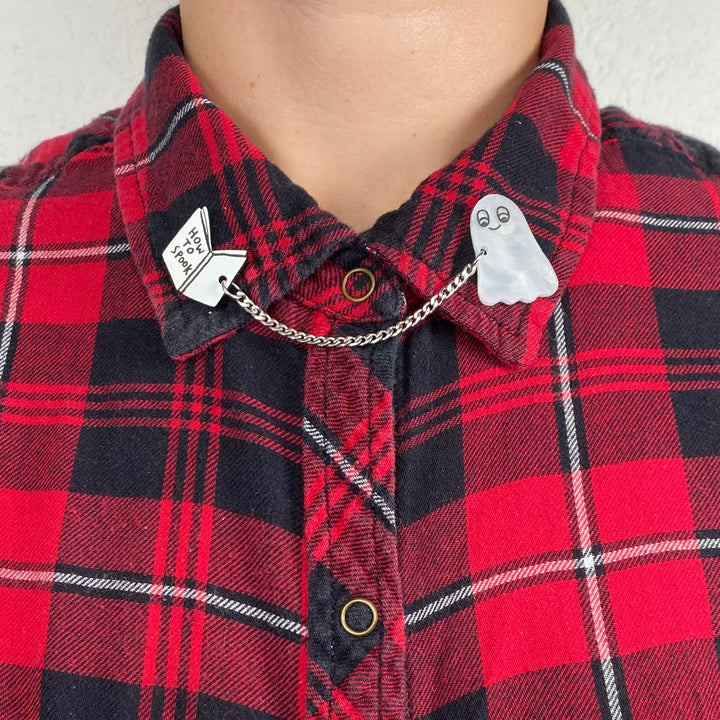 A person wears a red and black plaid shirt with our official licensed Gemma Correll Halloween-themed spooky ghost with 'How to' book collar clips.