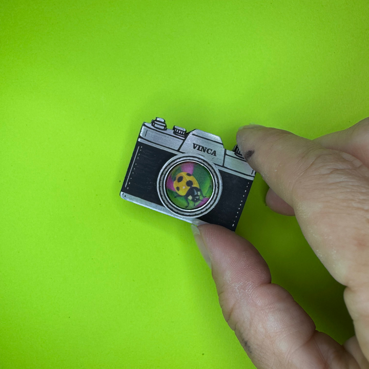 A hand holding a small camera-shaped hair clip with a colorful ladybug design in the center, set against a bright lime green background.