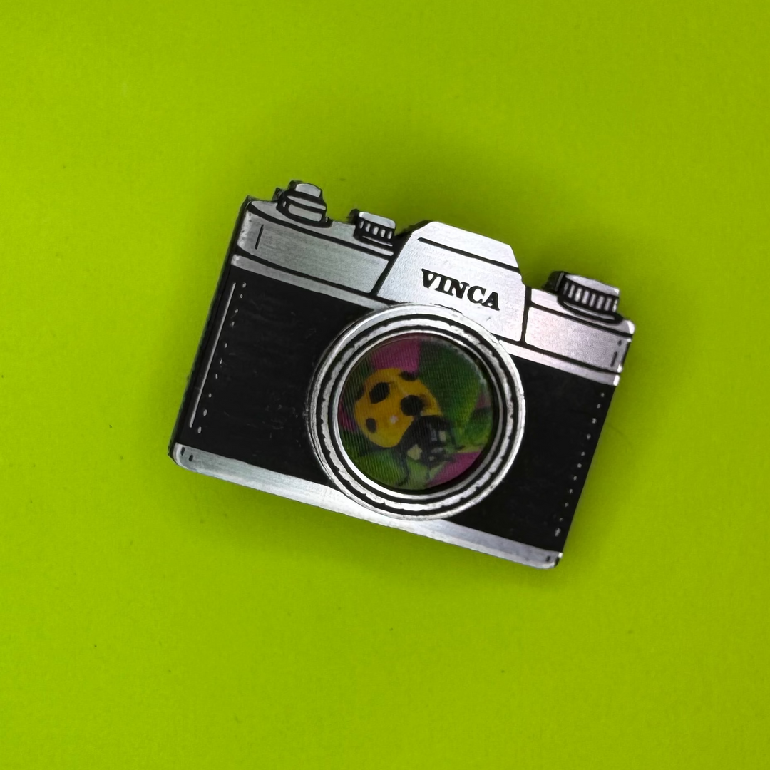 A vintage-style camera-shaped hair clip with 'VINCA' printed on the top, resting on a bright lime green background.