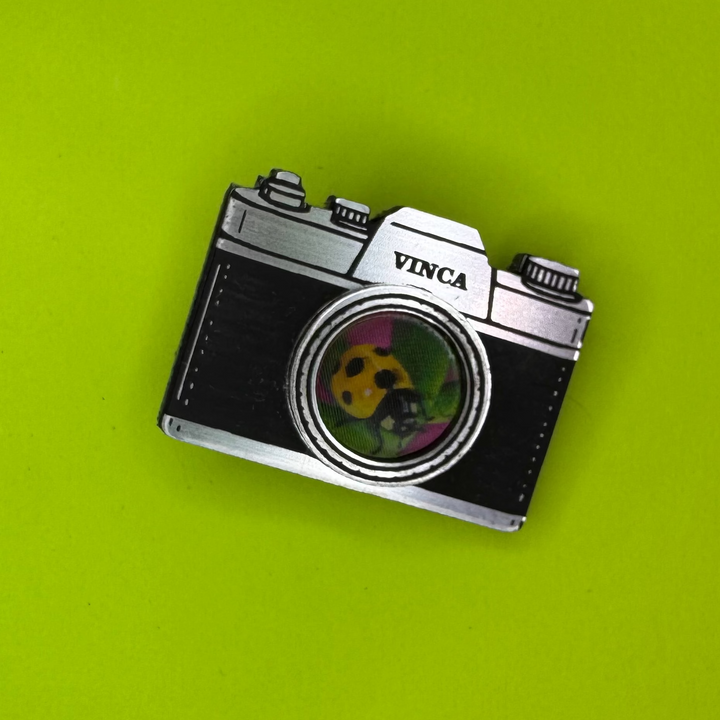 A vintage-style camera-shaped hair clip with 'VINCA' printed on the top, resting on a bright lime green background.