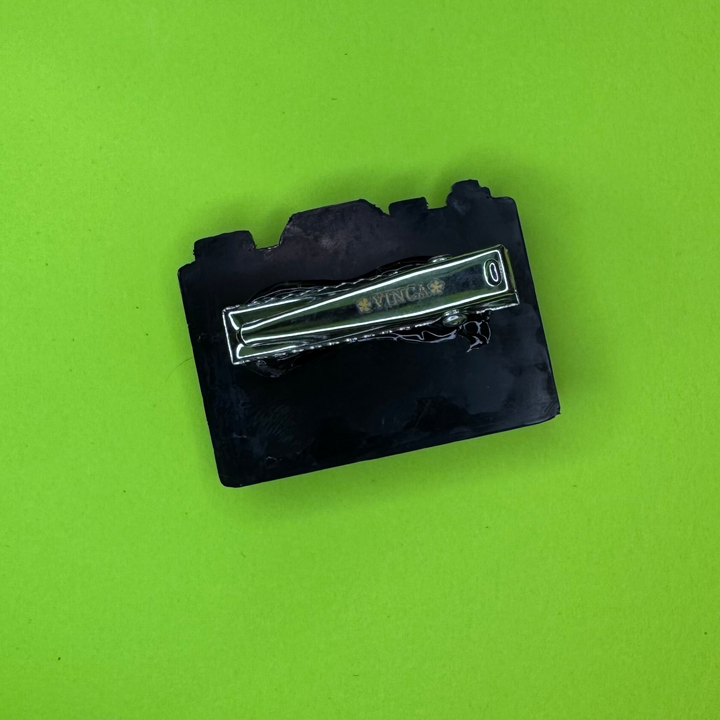 A small black rectangular camera shaped hairclip with a silver metallic clip across the center, photographed on a bright lime green background.