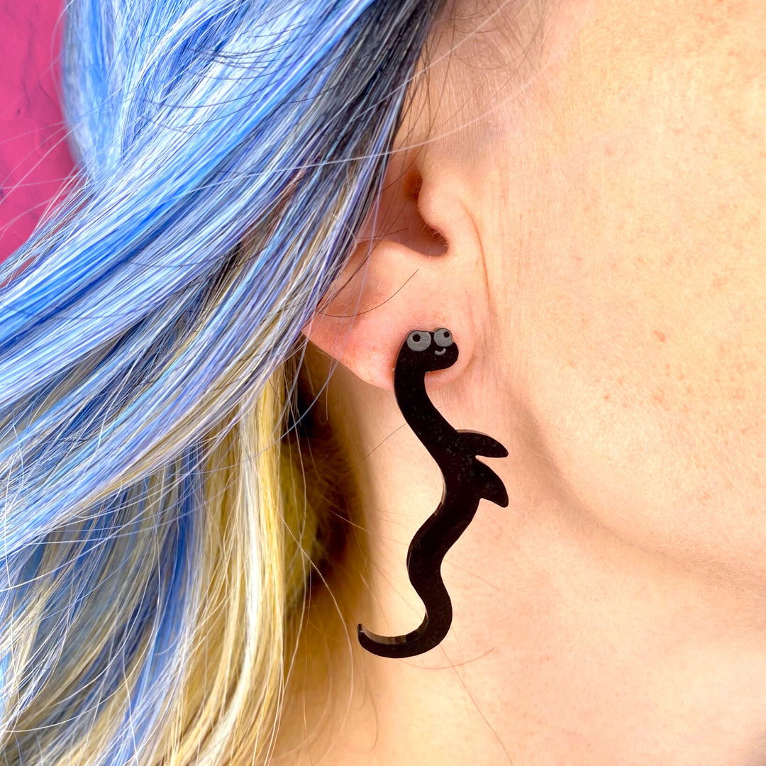 A model with blue hair wears our handmade Sarah's Scribbles Cryptid Club Nessie earring. 