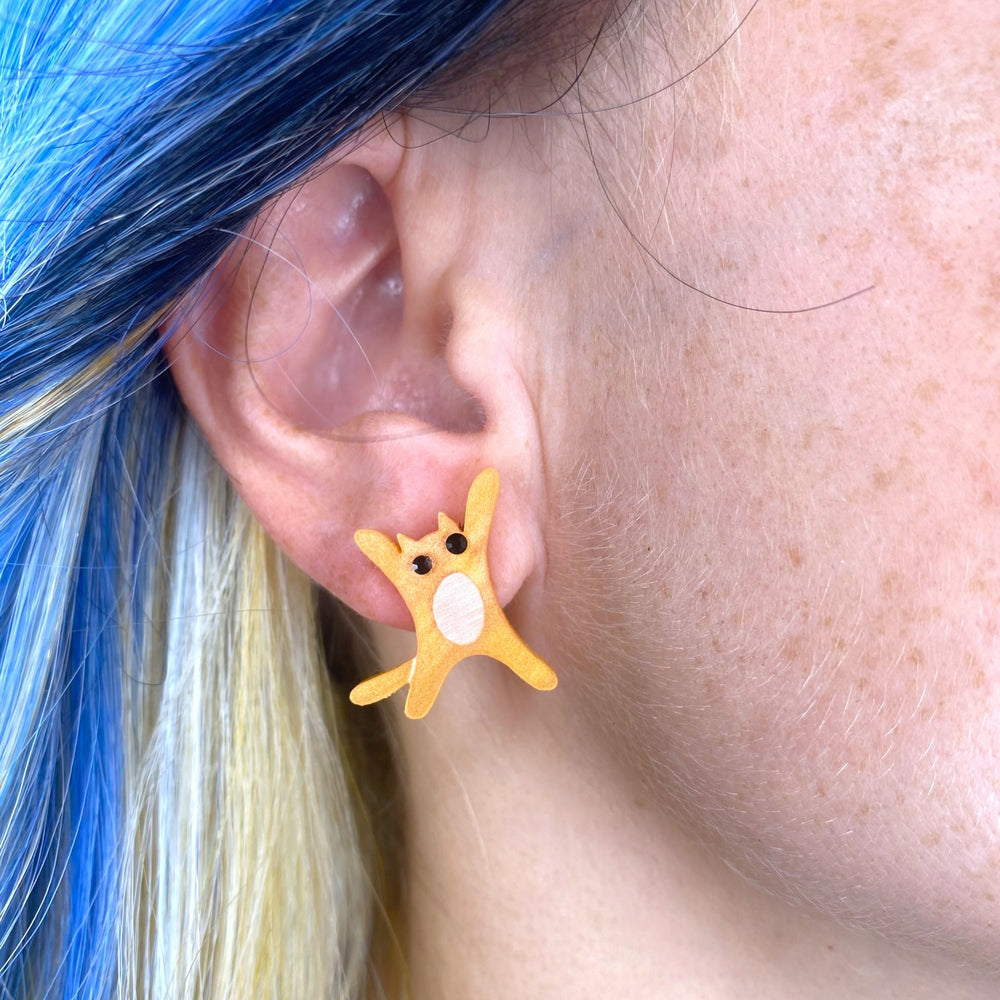 Person with blue and blonde hair wears a pearlescent orange cat earring illustrated by Sarah's Scribbles .