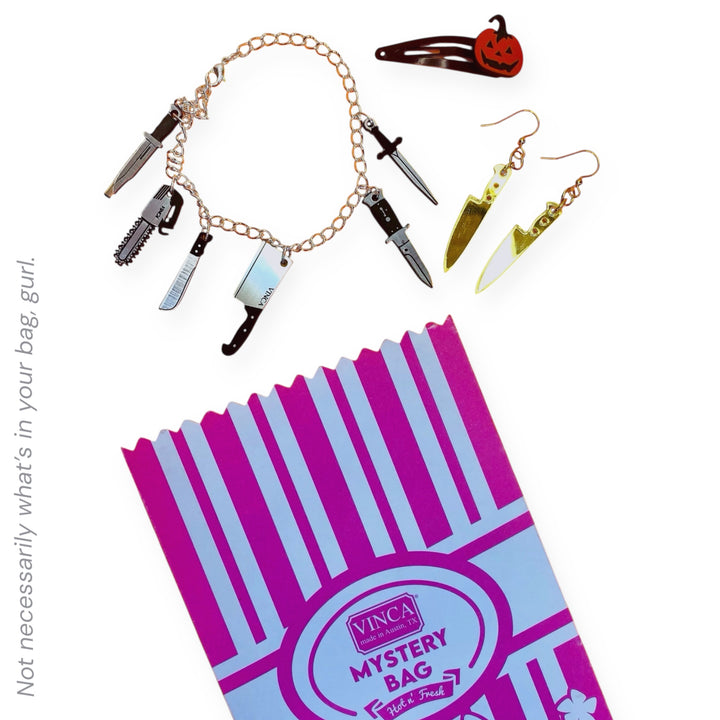 An sample of a Vinca Mystery Bag with DELUXE Halloween-themed jewelry including a six-piece charm bracelet, mirror gold chef's knife dangle earrings, and a Jack-o-lantern hair clip on a white background.