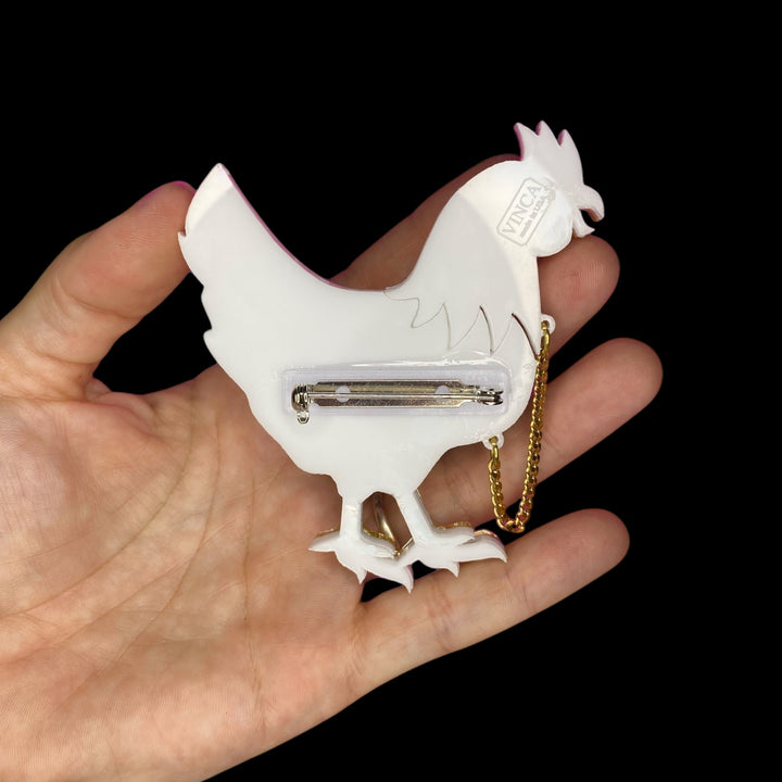 A hand holds the back side of our Chicken with its head cutoff brooch, which shows the pinback and branded back plate on a black background.