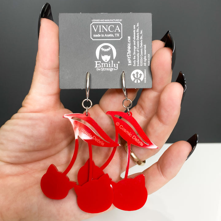 A model wearing black spider web nails holds our handmade officially licensed Emily the Strange red cat cherry hook dangle earrings against a white and black background. 