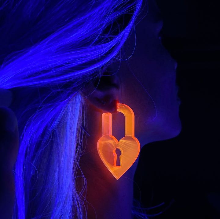 A woman wears a blacklight reactive neon pink heart-shaped lock earring  against a dark background.