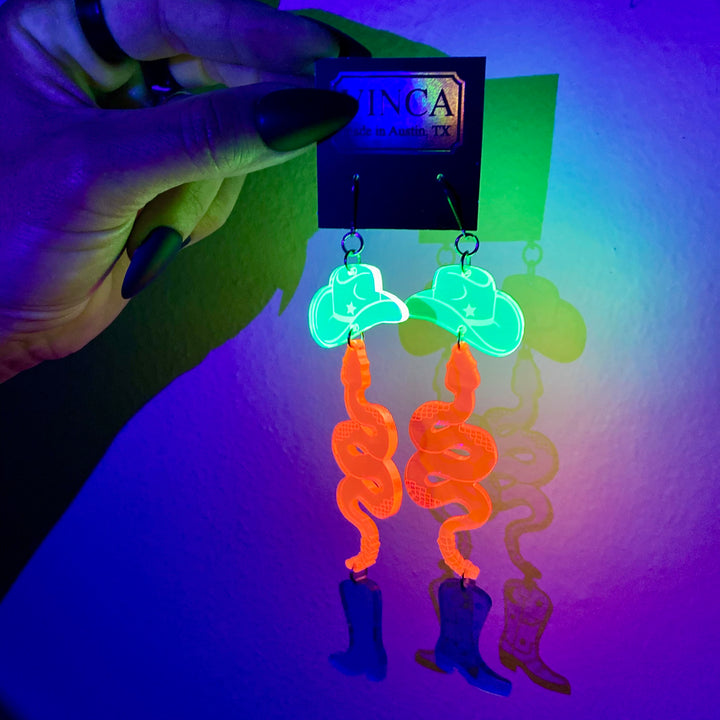 Glowing neon earrings with cowboy boot and hat design, held by a hand against a colorful background.