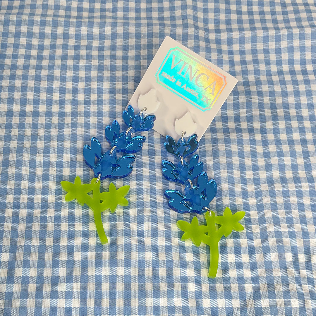 Extra large Texas Bluebonnet earrings attached to a white Vinca branded hangtag and on a gingham cloth.