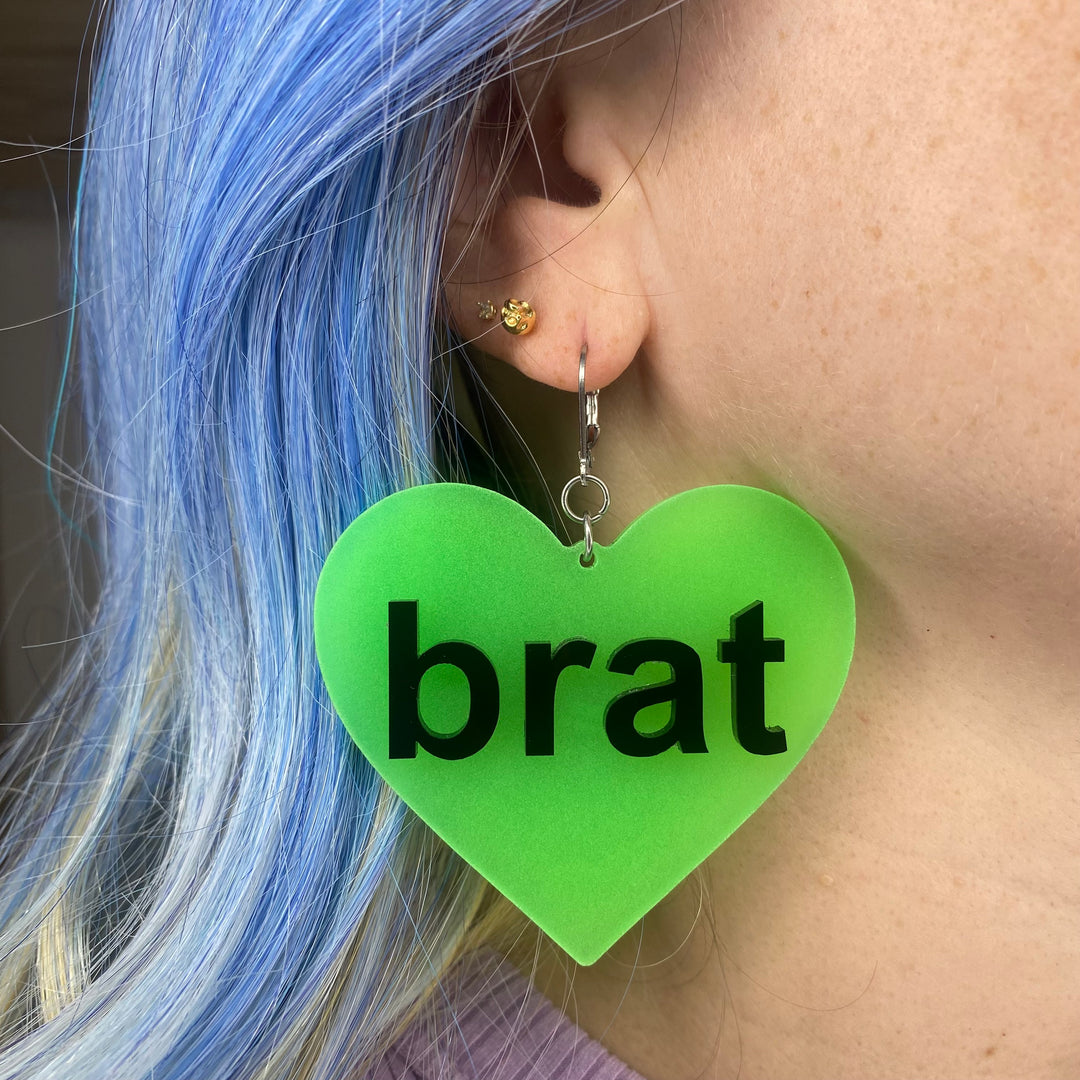 Super fun, extra large Brat Heart Dangle Earrings in glow-in-the-dark green are worn on the ear of a woman with bright blue hair.