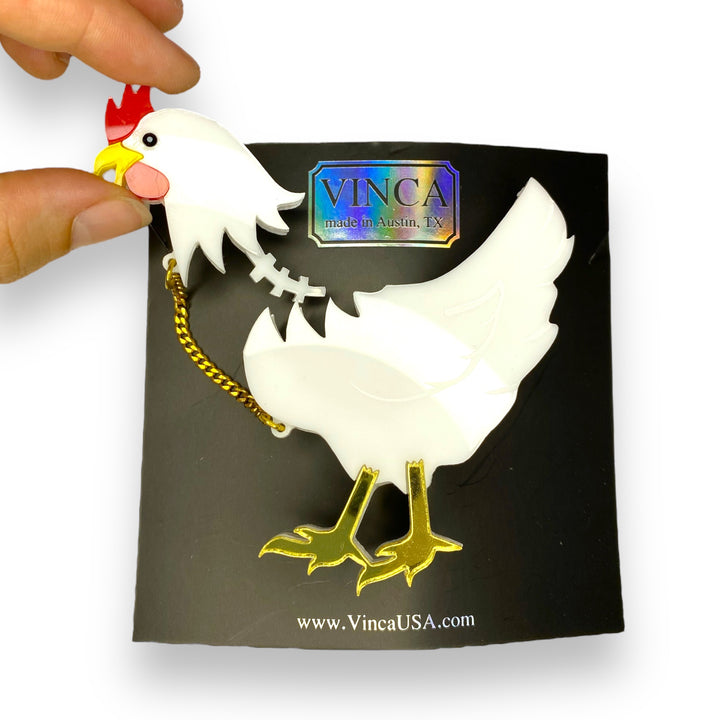 Fingers pulling off the head of a white chicken brooch that's attached by a gold plated stainless steel chain. Brooch is pinned to a black Vinca branded card set against a white backround.