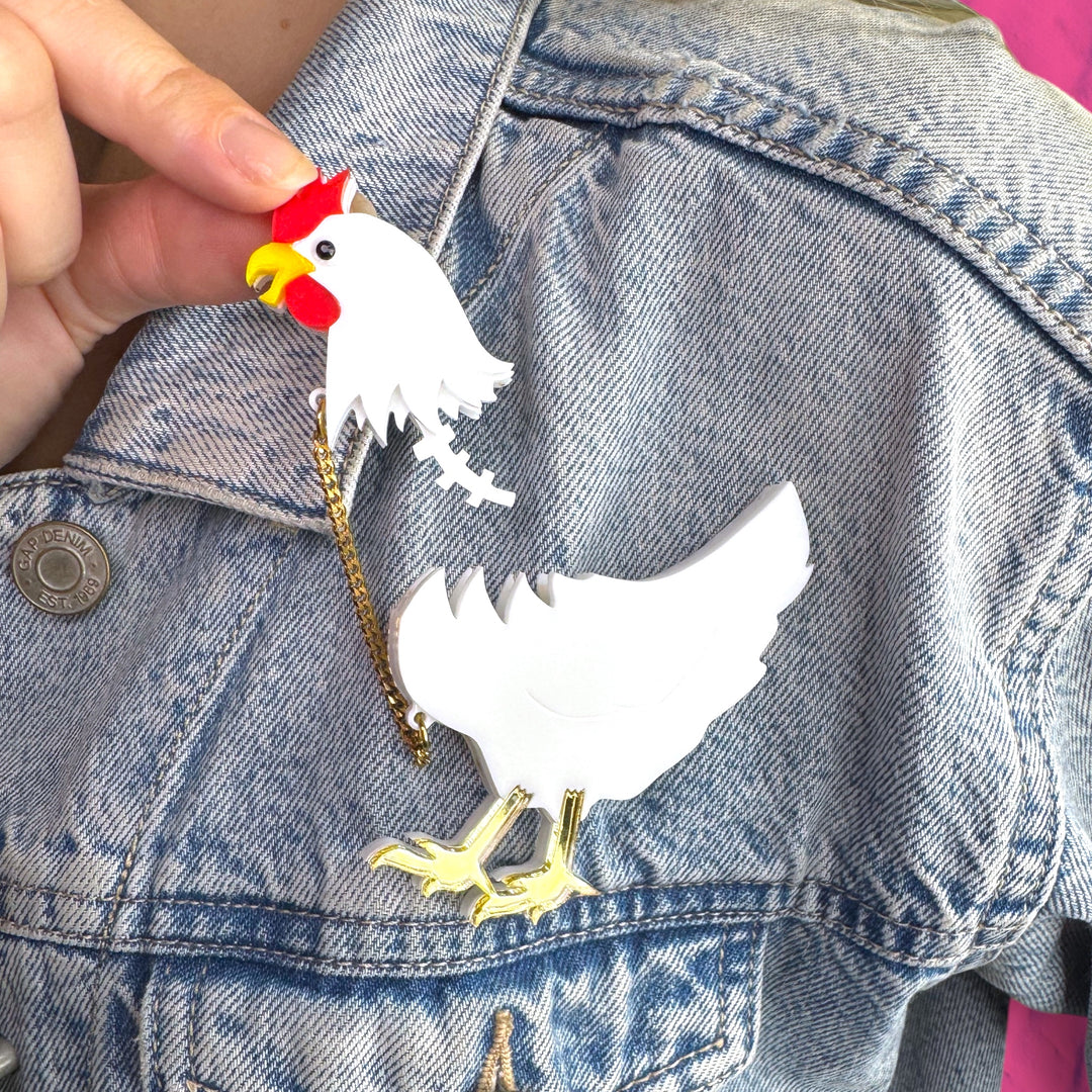 White chicken with detachable head brooch held together by a gold plated stainless steel chain  pinned to a jean jacket while its head is pinched between two fingers. 