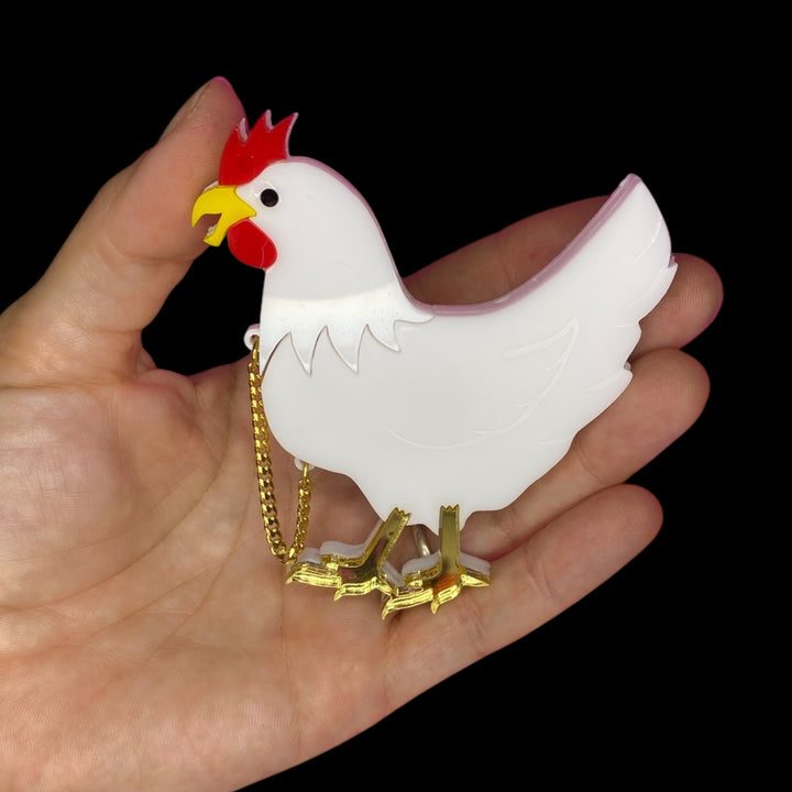 Chicken with its head cut off brooch is held in the palm of a hand set against a black backround.