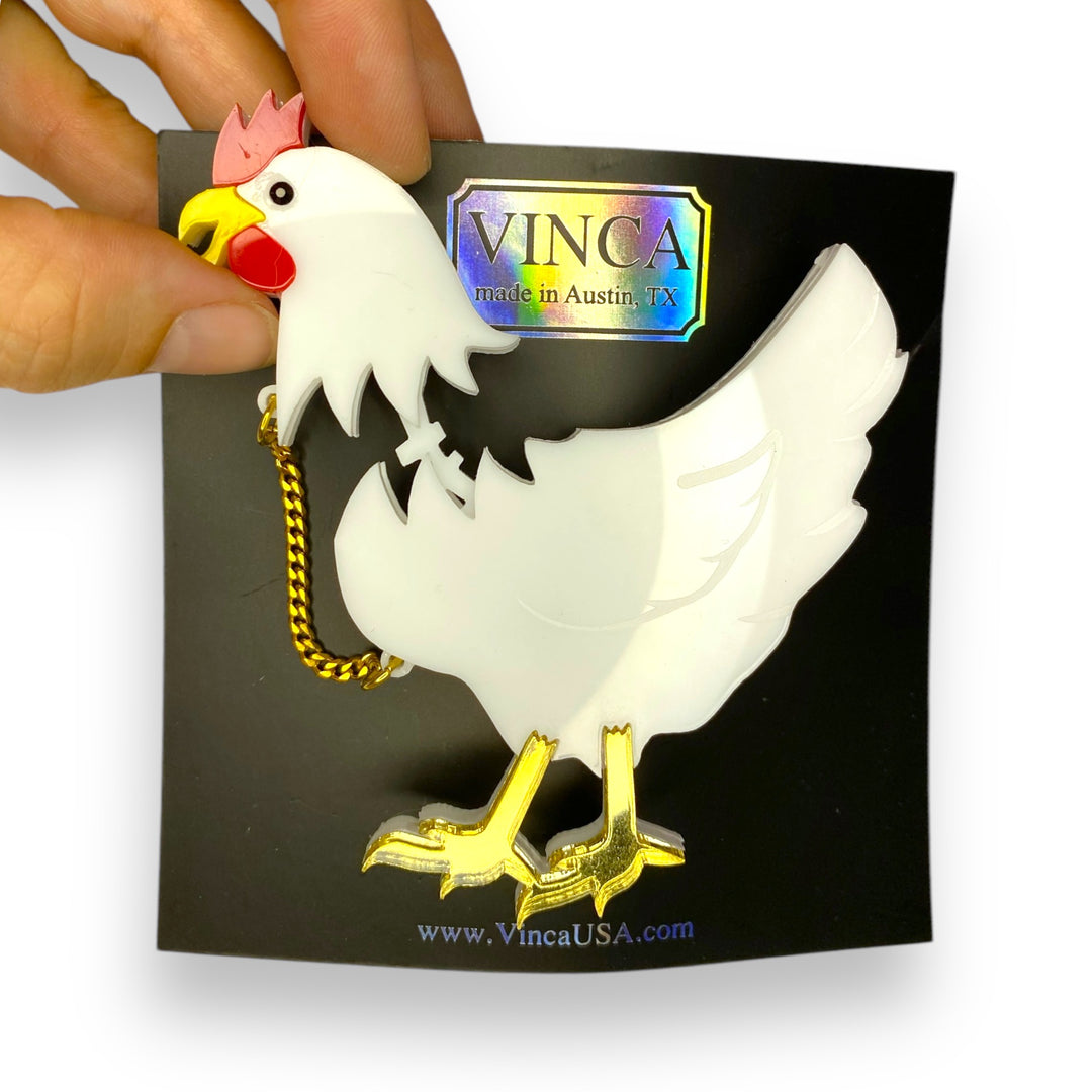 A hand is removing the head of our chicken brooch on a branded black Vinca card set against a white background. 