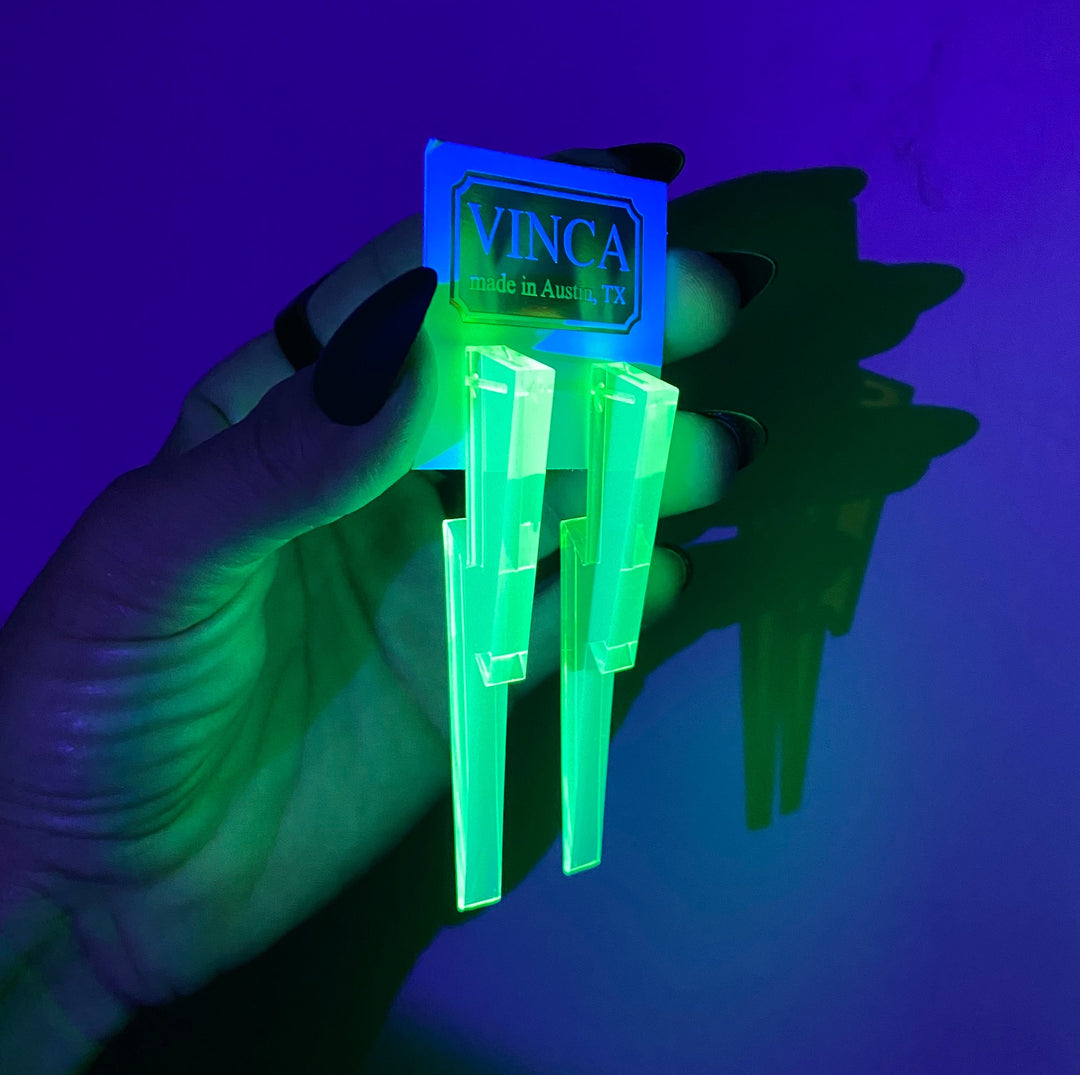 A hand holds blacklight reactive glowing green Chunky Lighting Bolt earrings against a dark background.