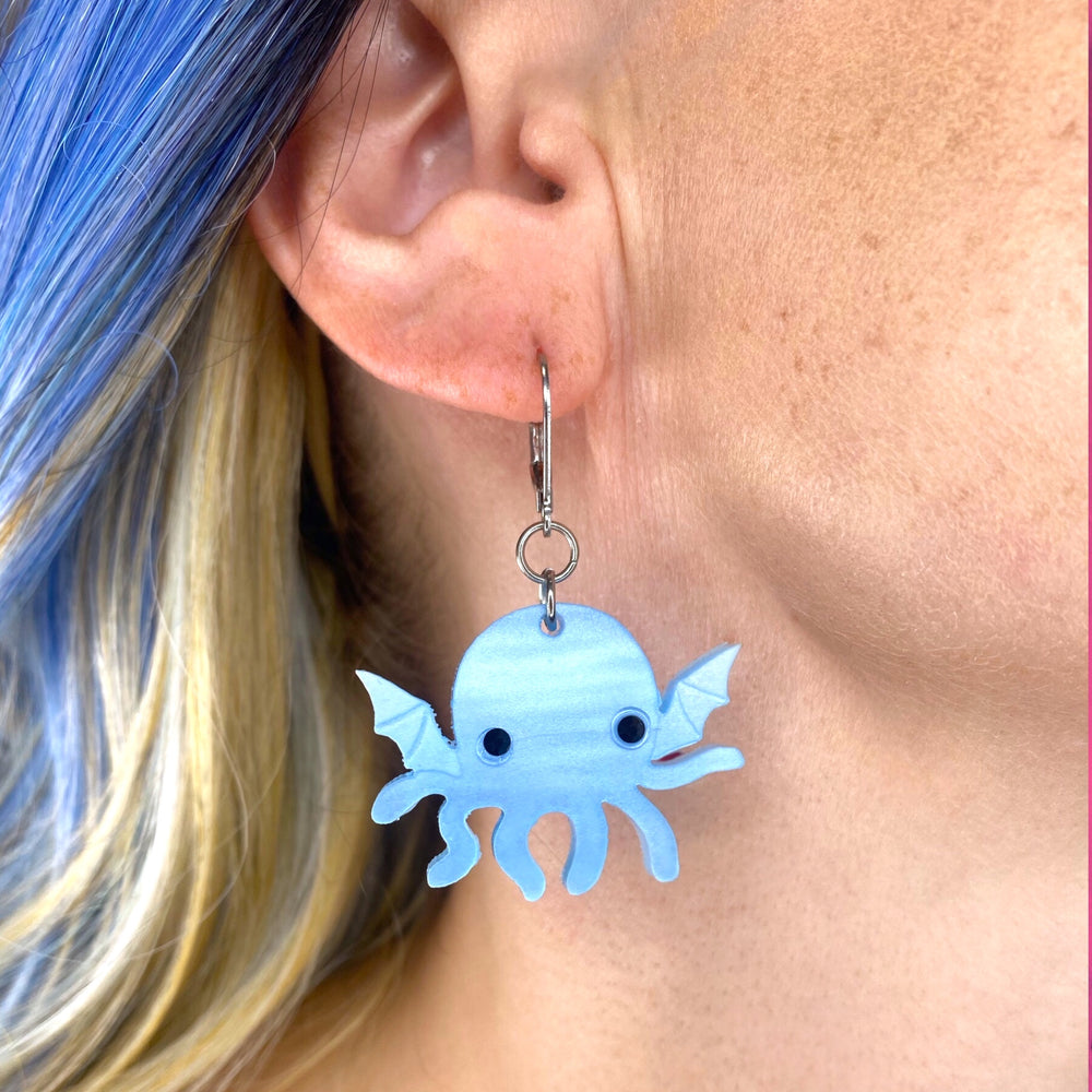 Our Sarah's Scribbles Cryptid Club Cthulhu danlge hook earrings are shown on a model with bright blue hair.