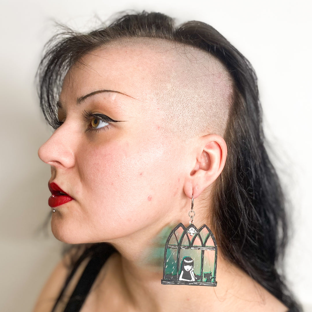 A pink rock model wears our officially licensed Emily the Strange soft goth iridescent window earrings against a white background.