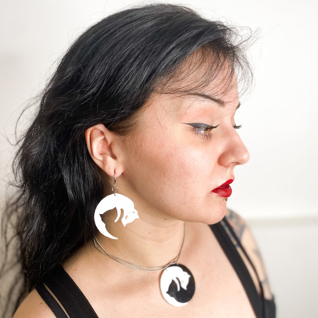 A model wears our officially licensed Emily the Strange Yin Yang Cat, black and white hook dangle earrings by a white background.