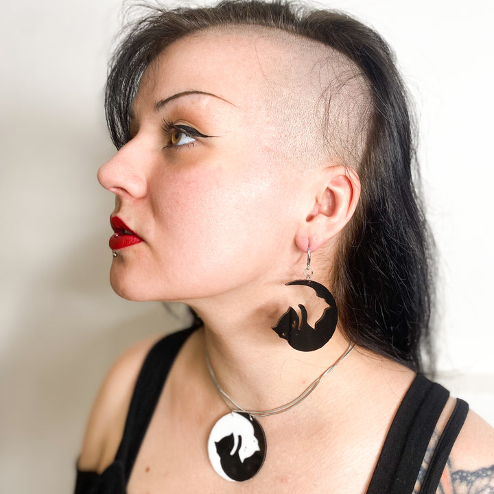 An edgy model wears our officially licensed Emily the Strange Yin Yang Cat, black and white hook dangle earrings by a white background.