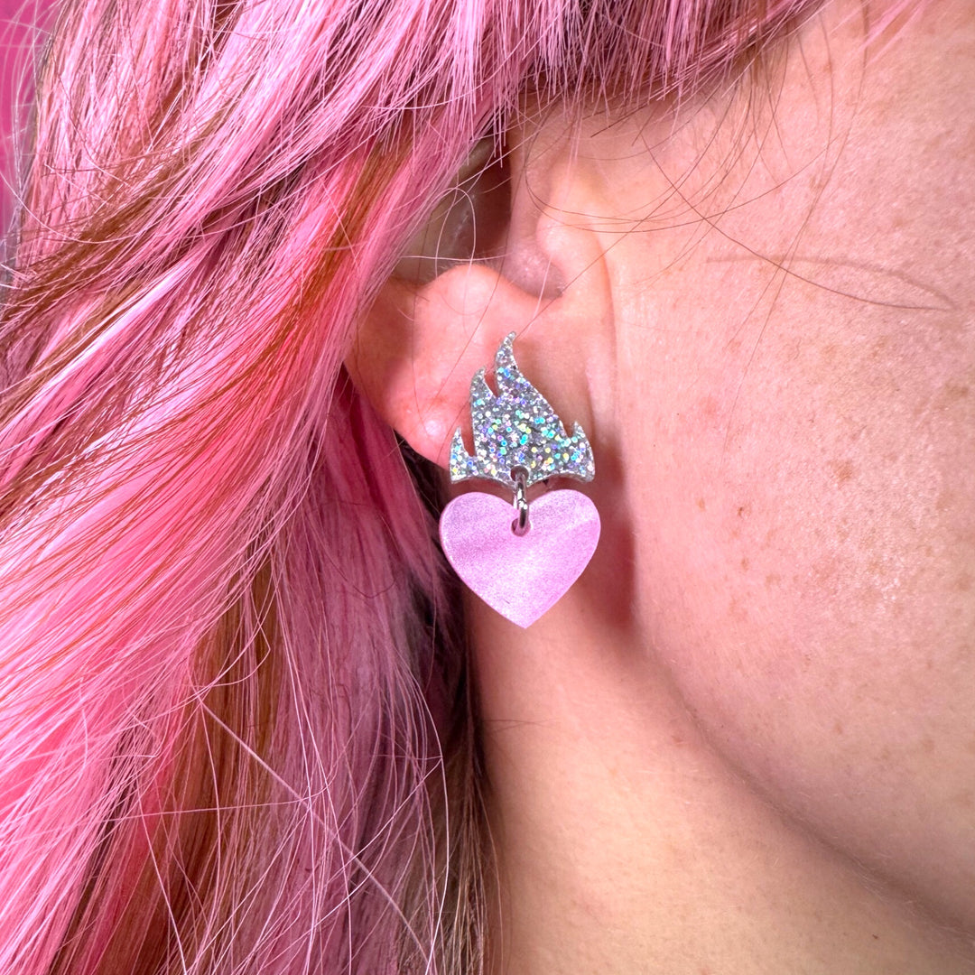 A model with pink hair wears our flaming heart earrings in pearl pink and glitter hologram.