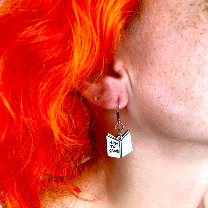 Person with bright orange hair wearing a spooky book dangle earring. 