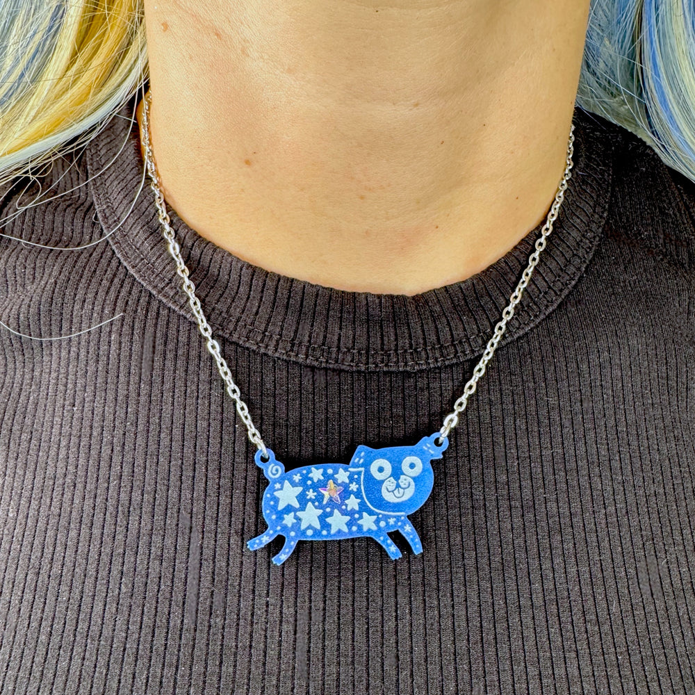 Person wearing a blue pug dog pendant on a chain necklace designed by Gemma Correll set against a dark ribbed sweater.