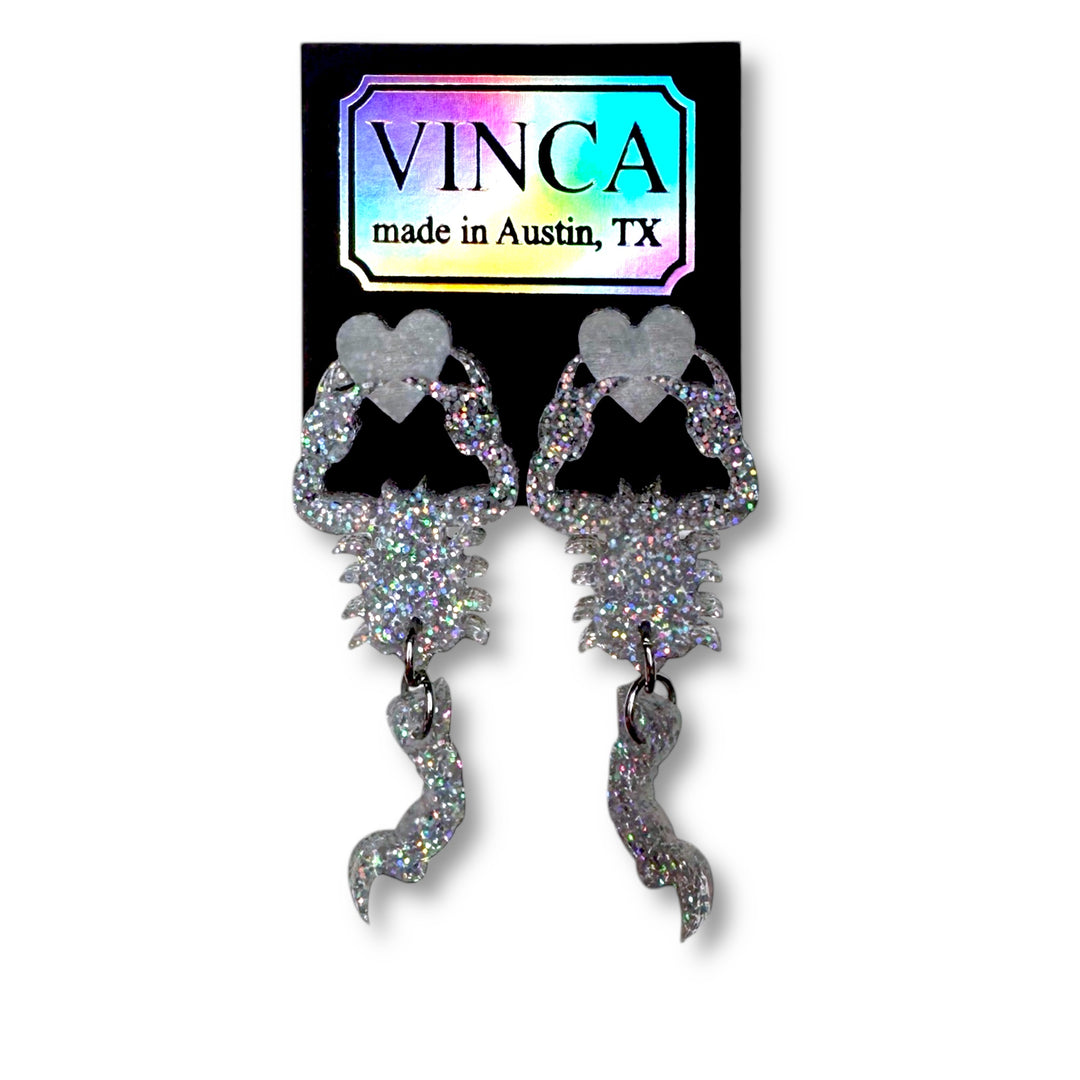 Glitter hologram scorpion holding a heart earrings on a Vinca branded card set against a white background.